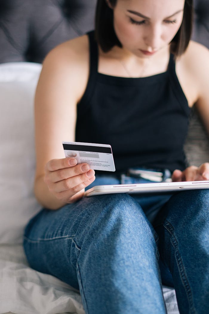 A woman in casual attire uses a credit card and tablet for online shopping while sitting on a bed.